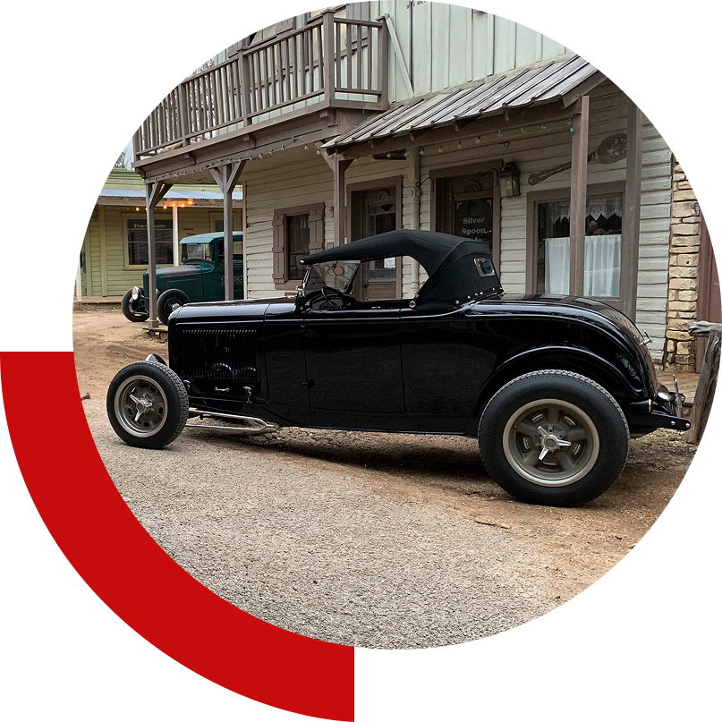 A black vintage convertible car is parked on a street in front of wooden buildings resembling an old western town.