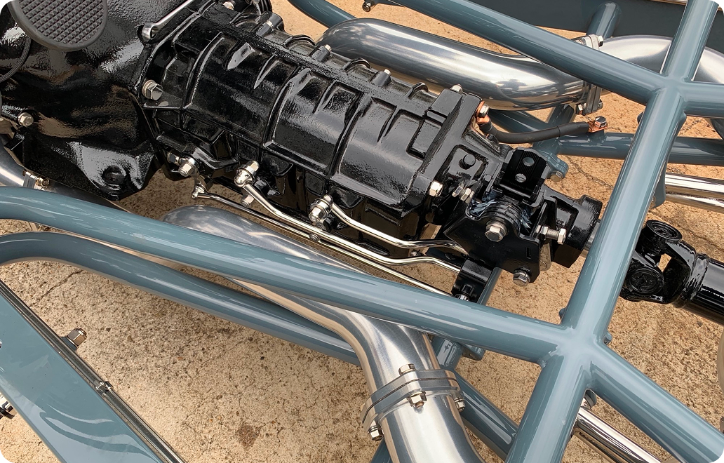Close-up of a black car transmission and silver exhaust pipes mounted within a gray tubular vehicle chassis on a concrete surface.