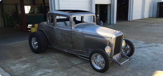 A vintage hot rod car with bare metal body, parked outside a garage with open bay doors on a concrete surface.