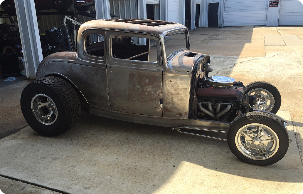 Unpainted vintage hot rod car parked on concrete, showing exposed metal body, large rear tires, and an exposed engine.